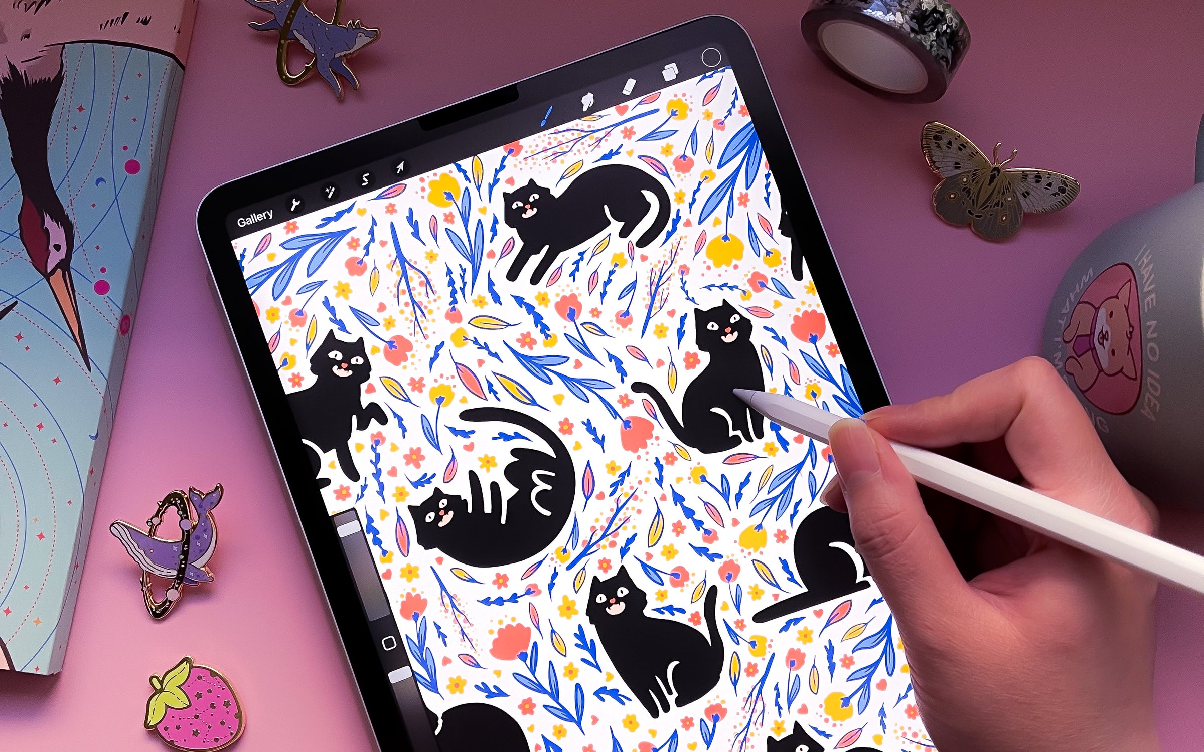 A photograph of an illustration featuring black cats and florals on an iPad surrounded by cute enamel pins.