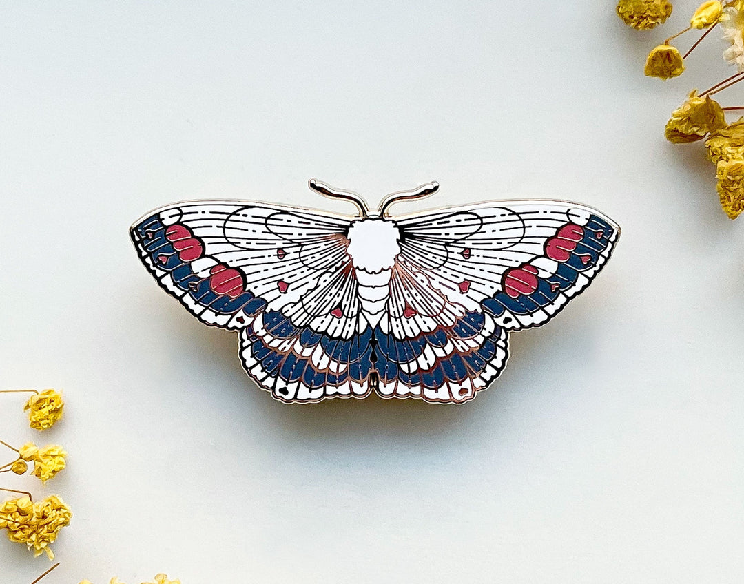 Decorative moth pin with red and gray patterns on a white hard enamel base. Pin is placed on a white background with yellow flowers.
